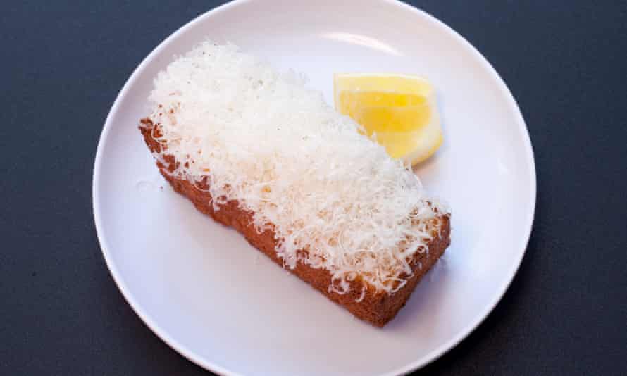 Un plato blanco redondo con un 'pastel' rectangular adornado con queso blanco rallado y una rodaja de limón en el lateral