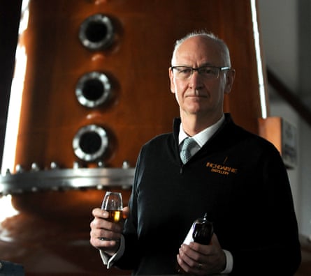 Ian Palmer frente a un alambique de cobre brillante, sosteniendo un vaso de whisky