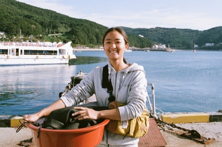 Ho-jin Shin, de 37 años, de pie sosteniendo un gran tazón para lavar platos en el borde de un puerto junto a un cuerpo de agua con la costa de la bahía en el medio.